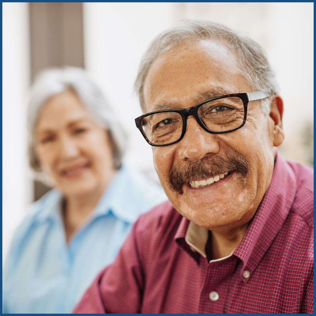 an older latino man smiling at the camera with a latina seated behind him.